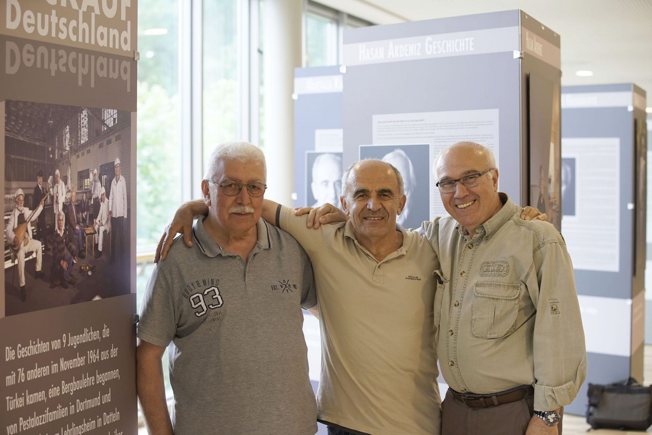 Drei ältere Männer stehen Arm in Arm zusammen. Im Hintergrund ist eine Ausstellung zu sehen.