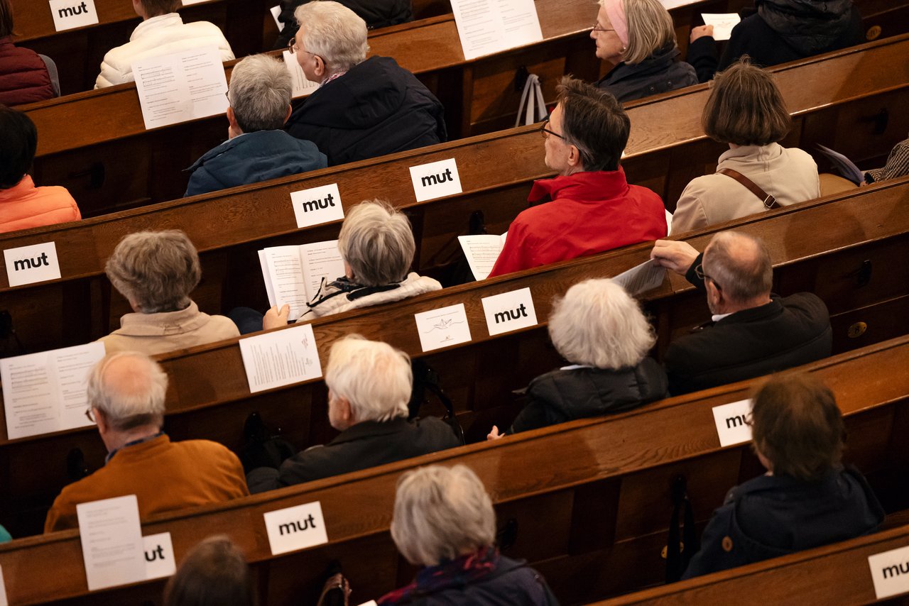Der Blick von oben: Menschen sitzen in Kirchenbänken. Vor ihnen liegen Zettel, auf denen Text steht. Auf kleineren steht das Wort Mut, klein geschrieben. Klick öffnet eine vergrößerte Ansicht.