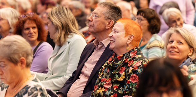 Ein Blick auf die bunt gemischte Zuhörerschaft. Im Mittelpunkt des Fotos sitzen eine ältere Frau mit auffällig rotem Haar und einer schwarzen Bluse mit Blumenmuster. Neben ihr eine älterer Mann mit kurzen Haaren. Er trägt eine Brille. Beide schauen aufmerksam lächelnd zur Bühne.