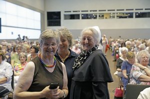 Drei ältere Frauen stehen in einem großem Saal. Im Hintergrund sitzen viele Menschen auf Stühlen. Eine Frau hat eine Kette mit bunten Holzperlen und hat ein Handy in der Hand. Eine andere Frau trägt eine Schwesterntracht. Die dritte Frau ist nur halb zu sehen.