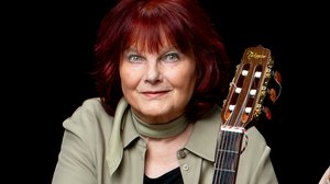 Porträtfoto JOANA: Eine ältere Frau mit roten Haaren steht hinter einer Gitarre.