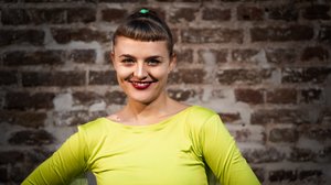 c_Beppo Lechenich Porträtfoto Hanna Held: Eine jüngere Frau mit kurzem Pony und rot geschminkten Lippen trägt ein neon gelbes Oberteil und hat die Hände in die Hüften gestützt.