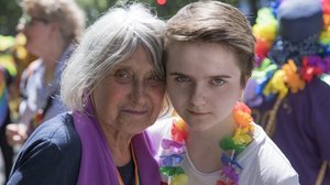 Eine ältere Frau mit grauen Haaren trägt ein Regenbogenband und einen lila Schaal um den Hals. Sie steht Kopf an Kopf mit einer jungen Person mit kurzen Haaren, die eine bunte Blumenkette um den Hals trägt.