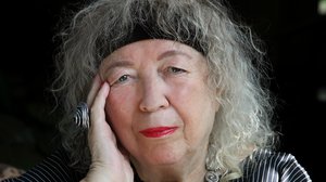 ©Kathrin Doepner Eine ältere Frau mit lockigen grauen Haaren. Sie trägt ein schwarzes Stirnband, ihre Lippen sind rot geschminkt. Sie stützt ihren Kopf auf ihre Hand, an der sie einen großen Ring trägt.
