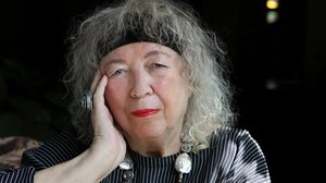 ©Kathrin Doepner Eine ältere Frau mit lockigen grauen Haaren. Sie trägt ein schwarzes Stirnband, ihre Lippen sind rot geschminkt. Sie stützt ihren Kopf auf ihre Hand, an der sie einen großen Ring trägt.