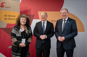 Drei Personen: Der Oberbürgeremeister Christian Specht, älterer Mann mit Halbglatze und Brille. Er trägt einen schwarzen Anzug, er hat seine Hände vor seinem Bauch gefaltet. Daneben der Bundeskanzler Olaf Scholz, älterer Mann mit Halbglatze. Er hat einen schwarzen Anzug an. Auch er hat die Hände gefaltet. Daneben ist die BAGSO-Vorsitzende Dr. Regina Görner, eine ältere Frau mit krausen Haaren. Sie alle schauen in die Kamera.