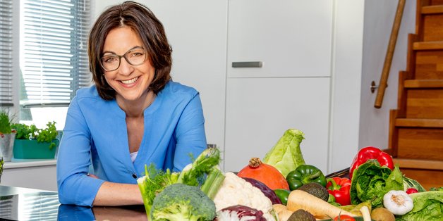 Eine Frau mit dunkelbraunem schulterlangem Haar steht an einen Küchentresen. Vor ihr stehen Körbe mit buntem Gemüse.