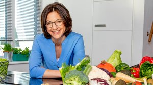 ©Claudia Timmann_NDR Eine Frau mit dunkelbraunem schulterlangem Haar steht an einen Küchentresen. Vor ihr stehen Körbe mit buntem Gemüse.
