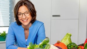 Eine Frau mit dunkelbraunem schulterlangem Haar steht an einen Küchentresen. Vor ihr stehen Körbe mit buntem Gemüse.