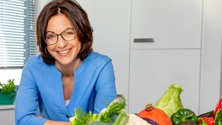 Eine Frau mit dunkelbraunem schulterlangem Haar steht an einen Küchentresen. Vor ihr stehen Körbe mit buntem Gemüse.