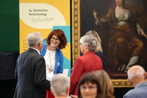 Drei Damen und ein Herr unterhalten sich lächeldn vor dem Banner des Deutschen Seniorentages.