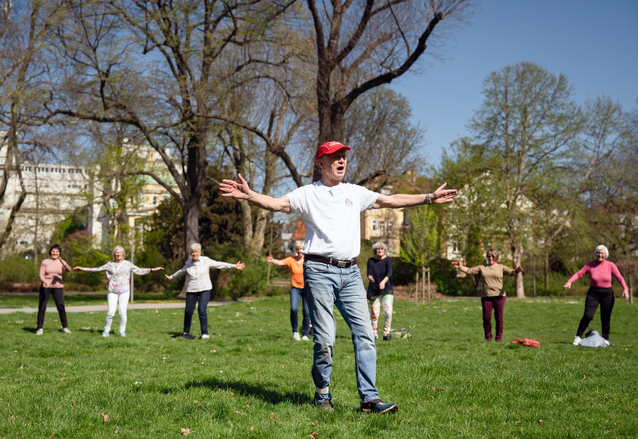 Ein älterer Mann in verwaschener Jeans, mit weißem T-Shirt, Turnschuhen und einer roten Kappe auf dem Kopf hat die Arme weit ausgebreitet und ruft etwas. Er steht in einem Park. Hinter ihm stehen Frauen, die Gymnastik machen. Klick öffnet eine vergrößerte Ansicht.