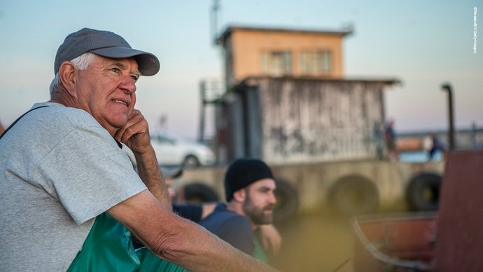 Ein Mann in Arbeitskleidung und Kappe sitzen am Kai eines Hafens und schaut in die Ferne. Im Hintergrund ein jüngerer Mann und ein Schuppen.