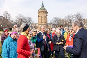 In einem Park stehen viele ältere Menschen. Im Hintergrund sieht man einen alten Turm.  In ihrer Mitte steht Franz Müntering, ein alter Mann. Vor Ihnen ein Mann mittleren Alters. Aus der Runde streckt eine Frau den Daumen nach oben. Alle lachen herzhaft.