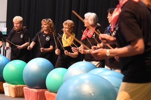 Eine Gruppe von schwarz gekleideten, älteren Frauen steht auf einer Bühne. Vor ihnen liegen jeweils ein blauer oder grüner großer Gymnastikball auf einem Gestell. Sie halten kurze Holzstöcke, die wie Trommelstöcke aussehen, in der Hand.