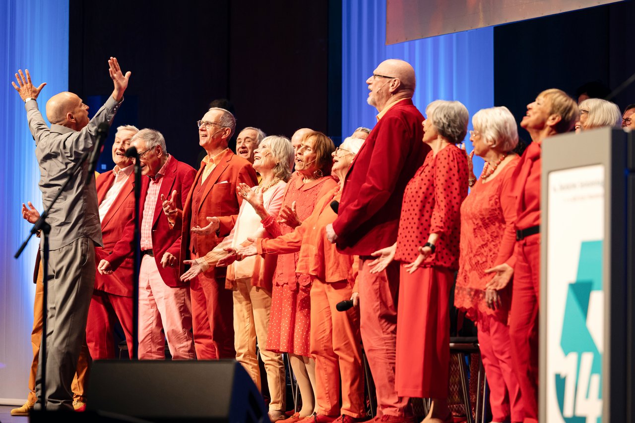 Ein Chor auf der Bühne: Alte Frauen und Männer, in Rottönen gekleidet singen gemeinsam. Vor ihnen steht der Chorleiter. Ein Mann mittleren Alters mit Glatze. Er is komplett in grau gekleidet. Er schaut den Chor an und hat seine Arme nach oben gestreckt. Klick öffnet eine vergrößerte Ansicht.