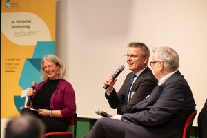 Zwei ältere Herren und eine Frau sitzen auf dem Podium und diskutieren.