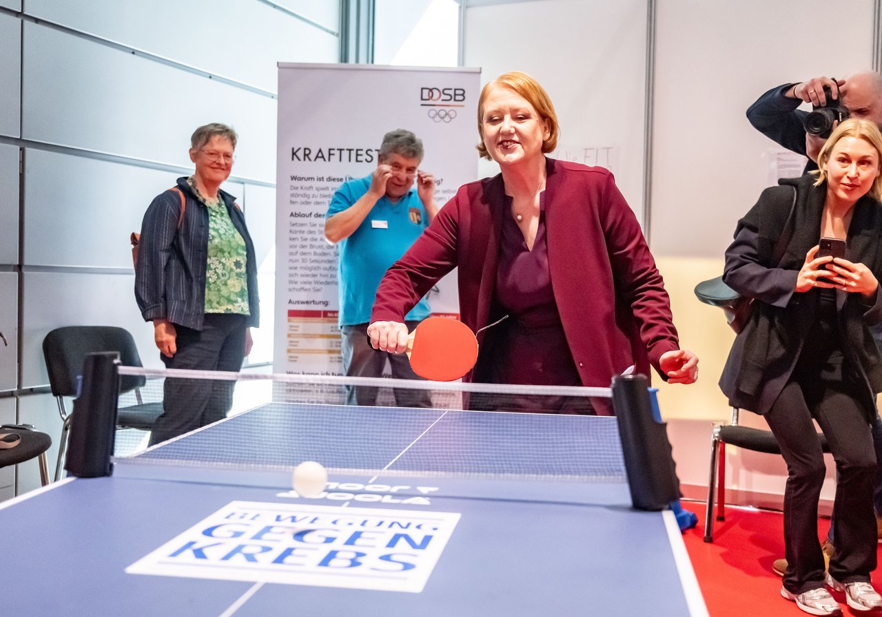 Im Vordergrund eine blaue Tischtennisplatte, mit der Aufschrift Bewegungen gegen Krebs. Die Seniorenministerin Lisa Paus spielt an der Platte Tischtennis. Sie trägt einen weinroten Blazer, darunter eine aubergine-farbene Bluse. Sie hat schulterlanges rötliches Haar. Um die Platte stehen Fotografen und andere Personen. Klick öffnet eine vergrößerte Ansicht.