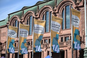 Vor dem Mannheimer Rosengarten, ein altes Gebäude aus rotem Sandstein, stehen Fahnenstangen. Daran sind gelbe Fahnen mit dem Logo und den Daten zum 14. Deutschen Seniorentag befestigt. Sie flattern im Wind..