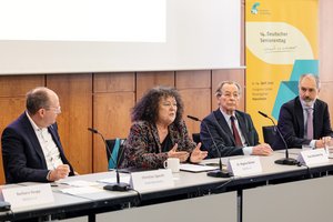Auf einem Podium sitzen eine Frau und drei Männer. Sie haben jeweils ein Mikrofon vor sich. Die Frau spricht gerade. 
