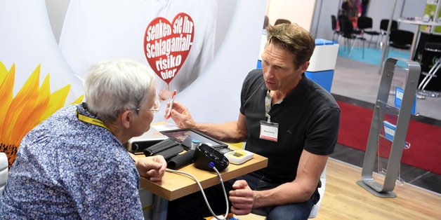 Ein Mann und eine ältere Frau sitzen an einem Messestand an einem kleinen Tisch. Vor ihnen liegt ein Tablet und ein Blutdruckmessgerät. Sie unterhalten sich. Im Hintergrund sieht man ein Plakat mit der Aufschrift "Senken Sie ihr Schlaganfallrisiko hier.