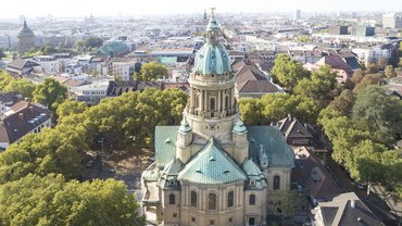 Die Mannheimer Christuskirche mit großer Kuppel und türkisen Dächern aus der Vogelperspektive. Die Kirche ist umringt von Bäumen.