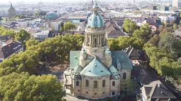 Die Mannheimer Christuskirche mit großer Kuppel und türkisen Dächern aus der Vogelperspektive. Die Kirche ist umringt von Bäumen.