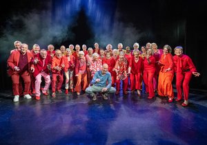 Eine großer Chor auf der Bühne. Alle sind älter und in verschiedenen Rottönen gekleidet. Vor ihnen sitzt eine Mann mit Glatze, der nicht bunt gekleidet ist in einer Hocke. Es ist der Chorleiter.