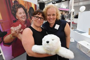 Zwei Frauen mittleren Alters stehen eng beieinander. Eine der beiden hat eine Eisbär aus Stoff auf dem Arm und hält ihn in die Kamera.