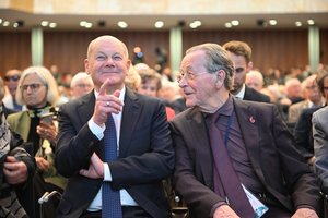 Bundeskanzler Olaf Scholz, ein älterer Mann mit Halbglatze und der ehemalige Minister Franz Müntefering, alter Mann im dunklem Anzug, sitzen nebeneinander. Olaf Scholz grinst und hebt einen Finger. Franz Müntefering schaut ihn an. Im Hintergrund ist Publikum zu sehen.
