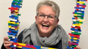 Eine ältere Frau mit Kurzhaarfrisur und Brille schaut durch eine Art Bilderrahmen, den sie in der Hand hat. Er ist aus bunten Legosteinen gebaut. Die Frau trägt einen grauen Schal und einen dunklen Pulli. Sie strahlt über das ganze Gesicht.