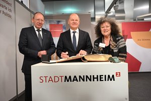 Hinter einem Stehpult, auf dem der Schriftzug "Stadt Mannheim" steht, stehen drei Personen. Der Oberbürgeremeister Christian Specht, der einen schwarzen Anzug trägt und die Hände vor sich gefaltet hält. Daneben der Bundeskanzler Olaf Scholz, älterer Mann mit schwarzem Anzug und Halbglatze. Er hält einen schwarzen Füller in der Hand. Auf dem Stehpult liegt ein großes, dickes Buch mit Goldschnitt, das goldene Buch der Stadt Mannheim. Daneben ist die BAGSO-Vorsitzende Dr. Regina Görner, eine ältere Frau mit krausen Haaren. Sie alle schauen in die Kamera.