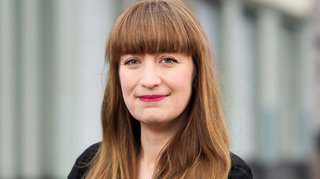 Portrait von Heidi Reichinnek: Junge Frau mit langem braunen Haar. Sie trägt ein weißes T-Shirt mit schwarzem Blazer. Sie steht mit verschränkten Armen seitlich zur Kamera.