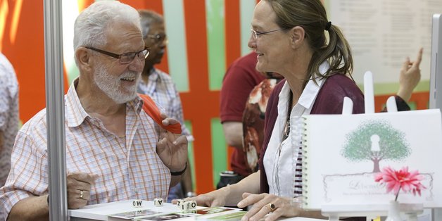 Ein älterer Mann mit grauem Bart und Brille steht an einem Messestand und spricht mit einer jüngeren Frau mit Zopf und Brille. Auf dem Stand liegen Würfel mit Buchstaben und Karten.