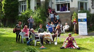 Ein großes Haus mit vielen Fenstern und rotem Dach. Auf der Wiese davor steht ein Tisch, um den viele verschiedene Personen sitzen, die unterschiedlich alt sind