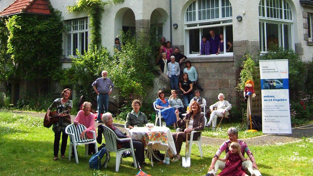 Ein großes Haus mit vielen Fenstern und rotem Dach. Auf der Wiese davor steht ein Tisch, um den viele verschiedene Personen sitzen, die unterschiedlich alt sind