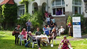 ©Freie Altenarbeit in Göttingen Ein großes Haus mit vielen Fenstern und rotem Dach. Auf der Wiese davor steht ein Tisch, um den viele verschiedene Personen sitzen, die unterschiedlich alt sind