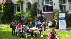 ©Freie Altenarbeit in Göttingen Ein großes Haus mit vielen Fenstern und rotem Dach. Auf der Wiese davor steht ein Tisch, um den viele verschiedene Personen sitzen, die unterschiedlich alt sind