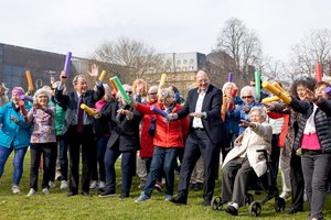 Eine Gruppe älterer Menschen machen auf einem Rasenstück gemeinsam Sport. Sie haben kurze, bunte Schwimmnudeln in der Hand. Sie strecken sie nach vorne und in die Höhe.