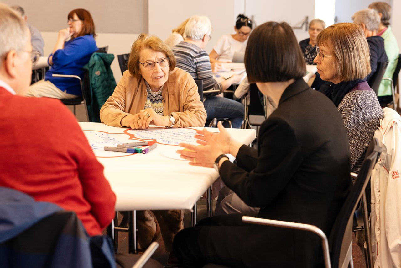Ältere Menschen sitzen um Arbeitstische, die mit Papier bedeckt sind und auf denen Stifte liegen. Im Fokus steht eine ältere Frau  mit beger Jacken, rötlichem Haar und Brille. Sie hört einer Frau, die ihr gegenüber sitzt, aufmerksam zu. Klick öffnet eine vergrößerte Ansicht.