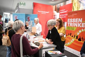 An einem Messestand stehen hinter der Theke Frauen, die diejenigen, die davar stehen beraten.