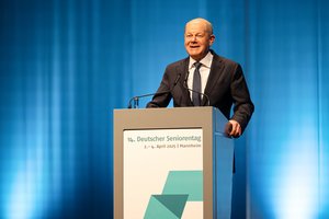 Bundeskanzler Olaf Scholz, ein älterer Mann in dunklem Anzug, weißem Hemd und Kravatte. Er steht an einem Rednerpult. Er lächelt ins Publikum.