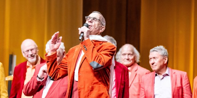 Auf der Bühne: Im Vordergrund ist ein alter Mann zu sehen. Im Anzug in Rottönen und einem weißen Schal um den Hals. Er singt mit geschlossenen Augen in ein Mikrofon. Hinter ihm ist ein Chor zu sehen. Auch in Rottönen gekleidet. Sie schauen auf ihn.