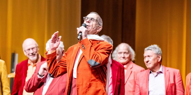 Auf der Bühne: Im Vordergrund ist ein alter Mann zu sehen. Im Anzug in Rottönen und einem weißen Schal um den Hals. Er singt mit geschlossenen Augen in ein Mikrofon. Hinter ihm ist ein Chor zu sehen. Auch in Rottönen gekleidet. Sie schauen auf ihn.