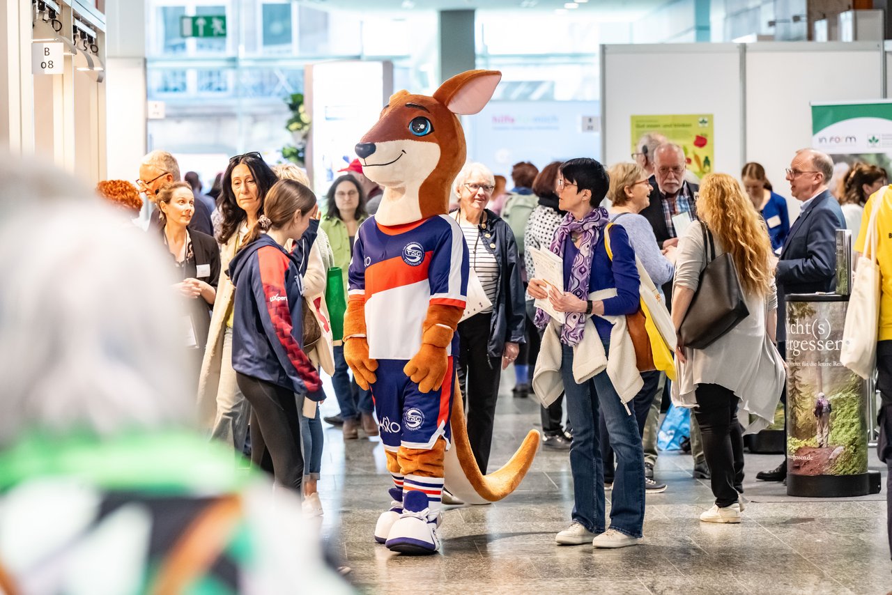 Ein Blick ins Messegeschehen. Viele Menschen stehen in unterschiedlichen Gruppen zusammen und unterhalten sich. Im Vordergrund eine Person in einem Kanguruhkostüm. Sie trägt eine Sporttrikot und eine kurze Sporthose in blau-orange. Die Figur überragt alle. Klick öffnet eine vergrößerte Ansicht.