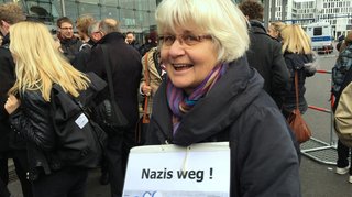 Eine Frau mit weißen kinnlangen Haaren und Brille trägt ein Schild auf dem „Nazis weg!“ steht.