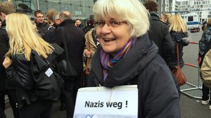 Eine Frau mit weißen kinnlangen Haaren und Brille trägt ein Schild auf dem „Nazis weg!“ steht.