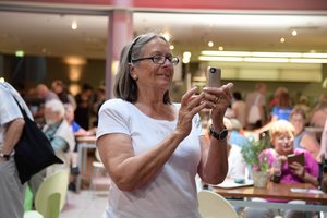 Eine ältere Frau steht in einem Saal. Im Hintergrund sitzen Menschen an Tischen. Die Frau hält ein Handy in der Hand und schaut darauf.