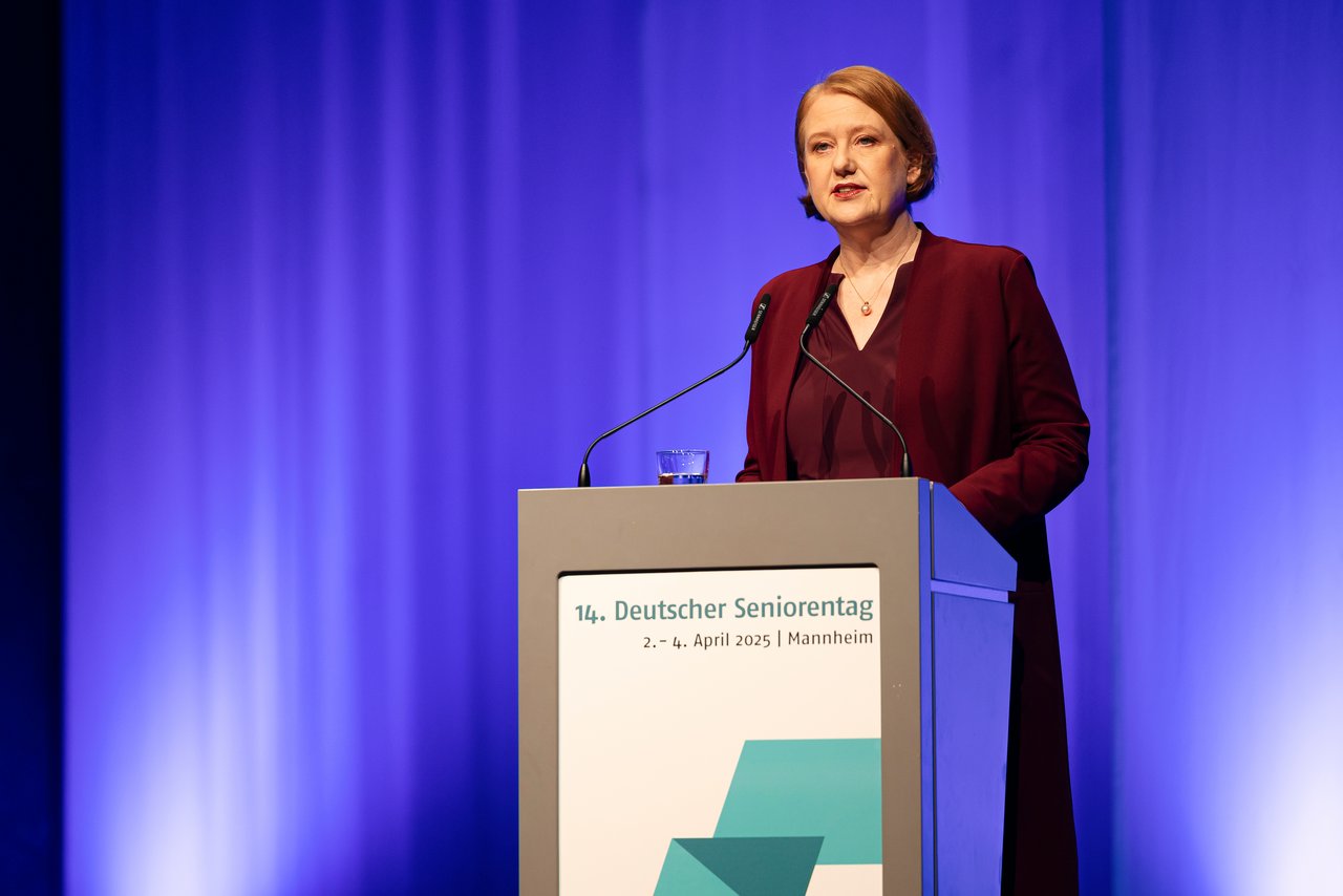 Die Seniorenministerin Lisa Paus, eine Frau mittleren Alters mit rötlichem, schulterlangen Haaren. Sie trägt einen dunkelroten Blazer und eine Bluse. Sie steht am Rednerpult und spricht ins Publikum. Klick öffnet eine vergrößerte Ansicht.