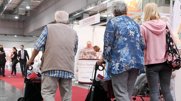 Ein älterer Mann und zwei Frauen mit Rollatoren - nur von hinten zu sehen -  laufen durch die Messehallen auf dem Deutschen Seniorentag in Dortmund.